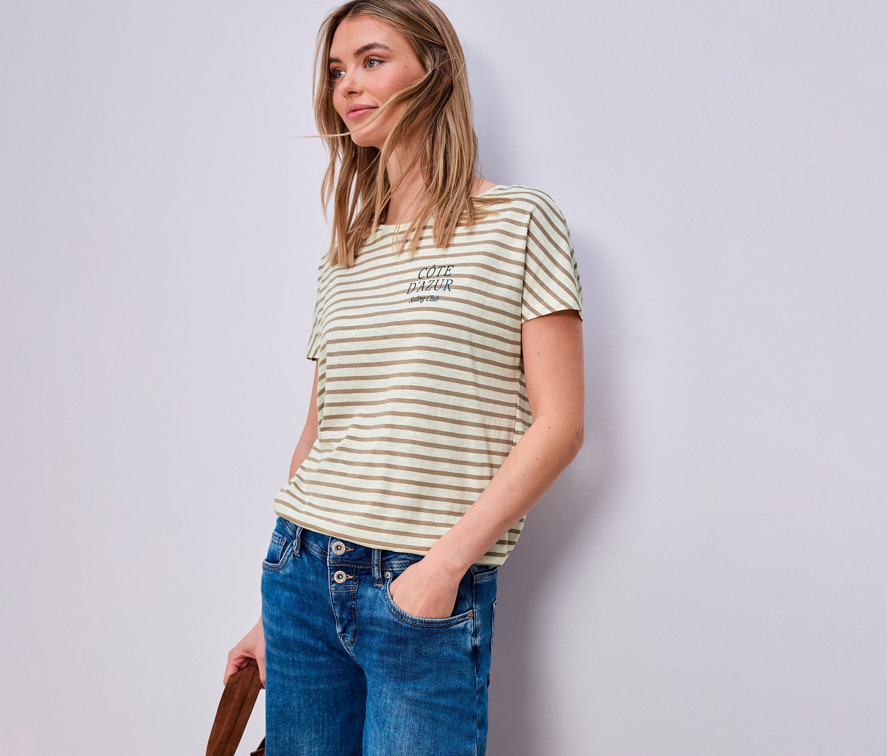 Frau mit Jeans und beige-weißem Street One T-Shirt. Sie hat eine Hand in der Hosentasche und hält eine braune Tasche.