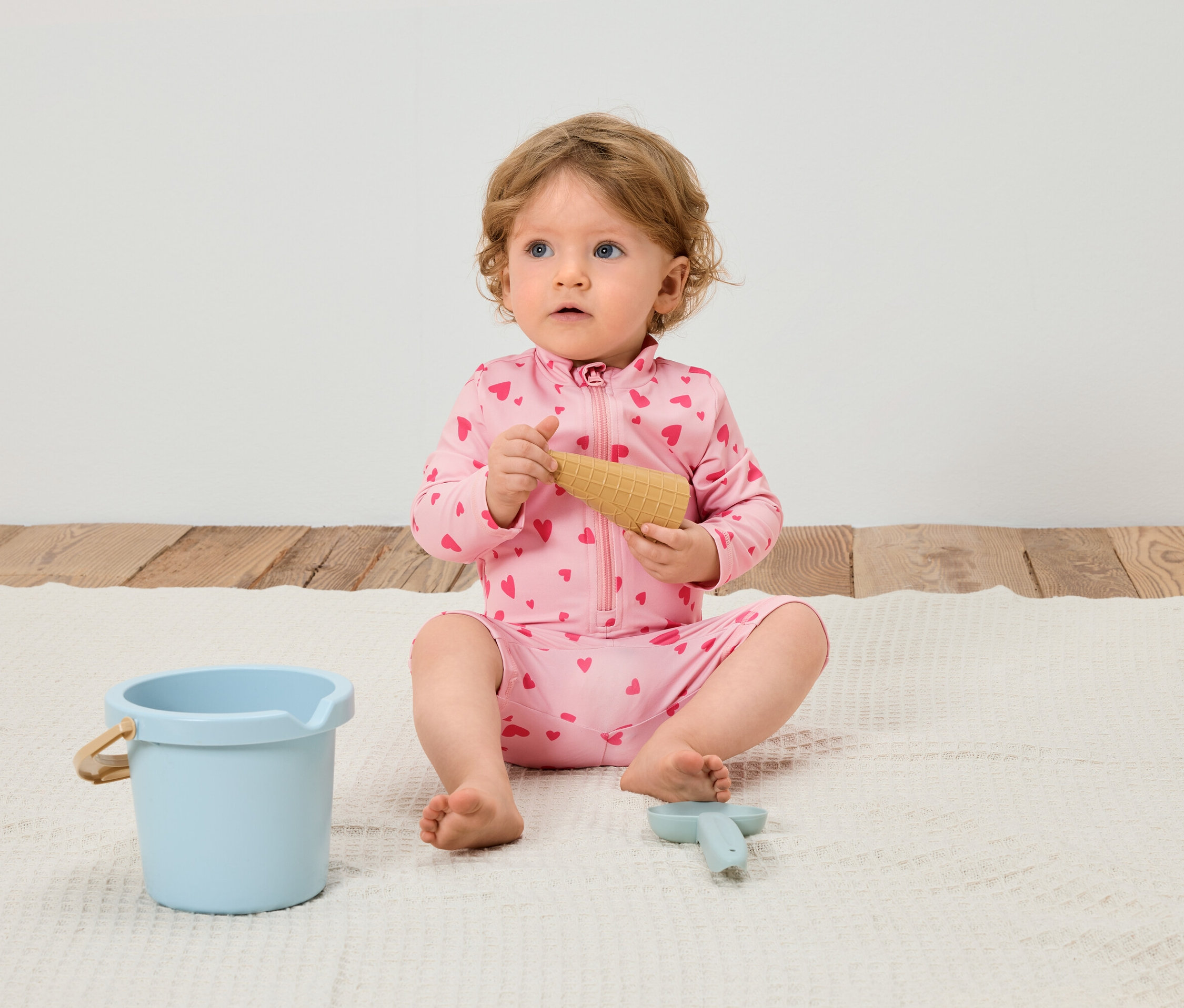 Ein Baby mit blonden Locken sitzt auf einer weißen Decke und spielt mit einem Spielzeugeis, einem blauen Eimer und einer blauen Schaufel.