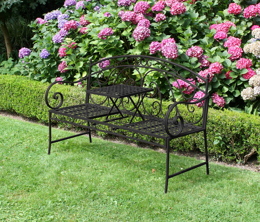 Eine schwarze Metallbank mit integriertem Tisch steht auf einer grünen Wiese vor einem Busch mit rosa und lila Hortensien.