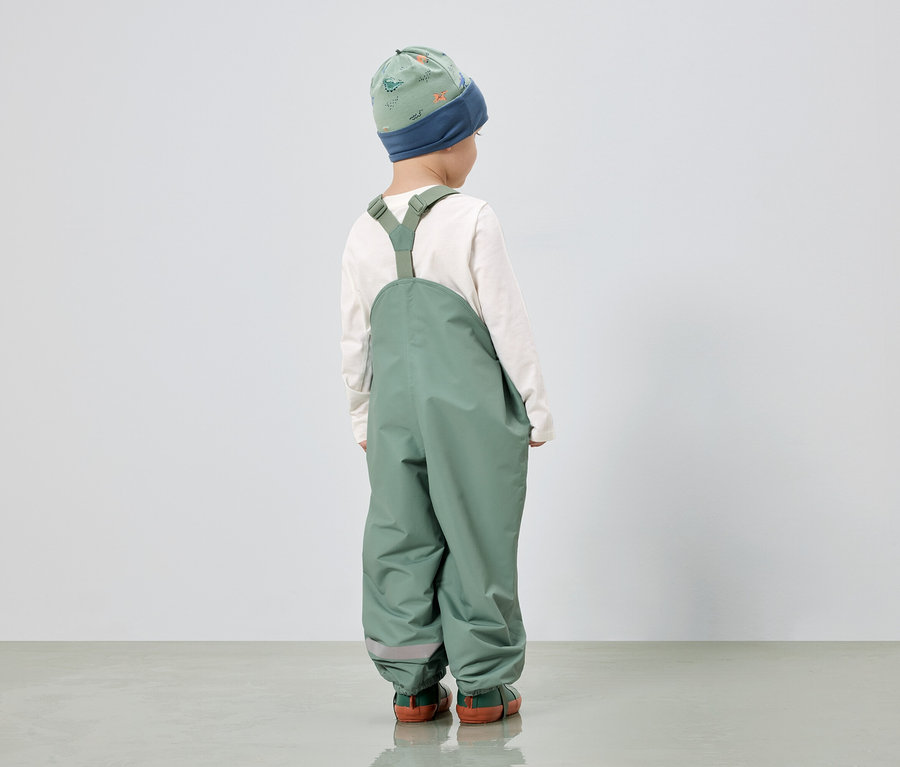 Kind von hinten, trägt grüne Latzhose, ein langärmliges, weißes Oberteil und eine grüne Mütze mit Dinosaurier-Muster.