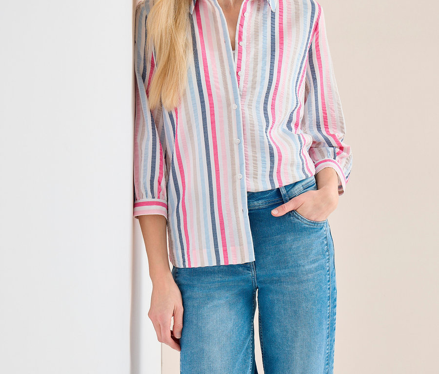 Blonde Frau mit hellblauer Kappe, gestreifter CECIL Hemdbluse in Pink, Blau, Grau und Weiß, und blauen Jeans, die an einer Wand lehnt und lächelt.