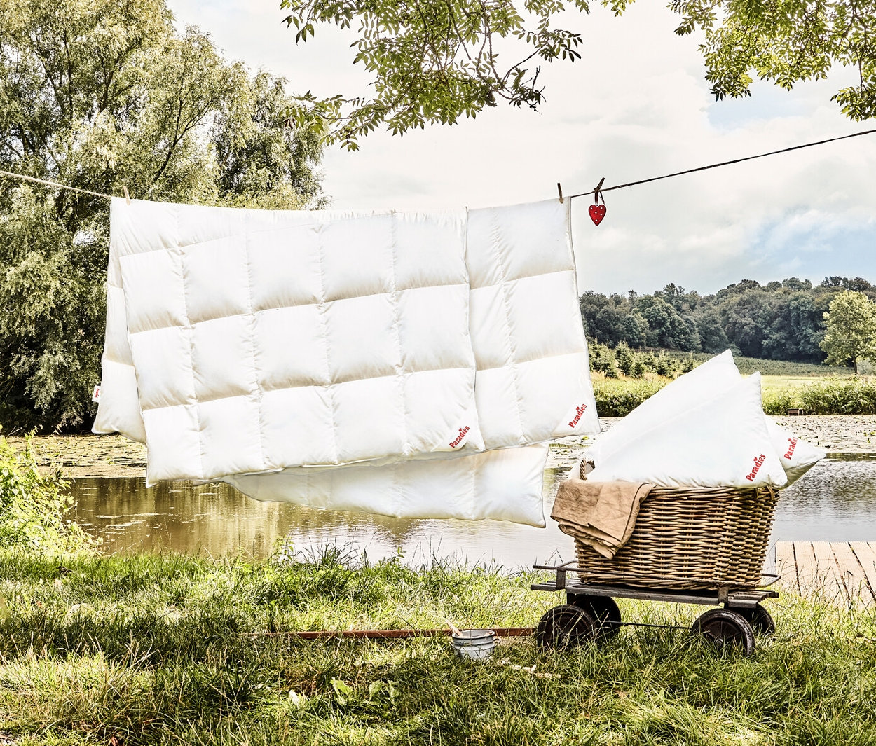 Eine Paradies Ganzjahres-Daunendecke »Hoheluft« in Normalgröße hängt draußen an einer Wäscheleine, während eine weitere Decke und Kissen in einem Korbwagen liegen.