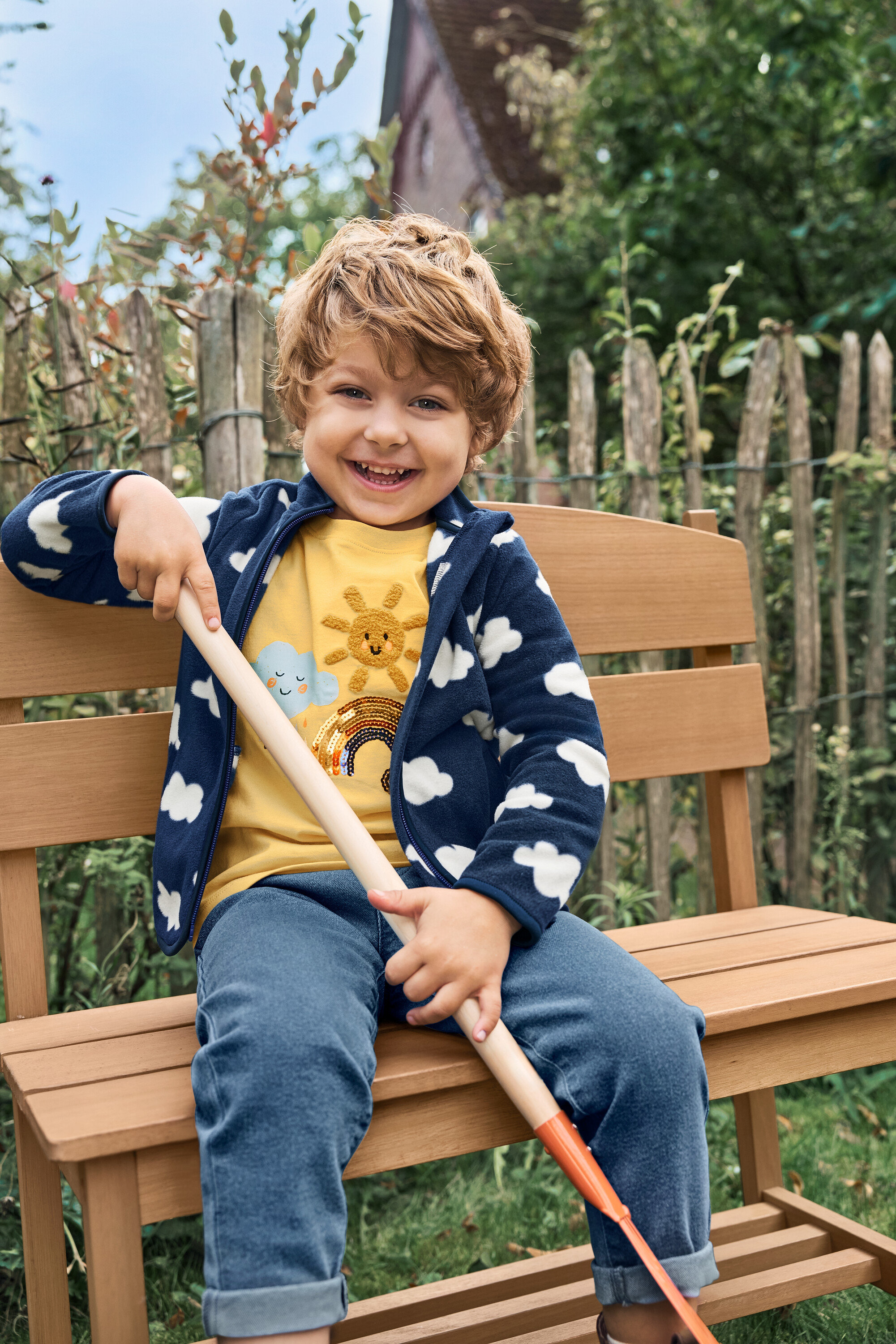Ein lächelnder Junge sitzt auf einer kleinen Gartenbank mit Ablage und hält eine Schaufel.