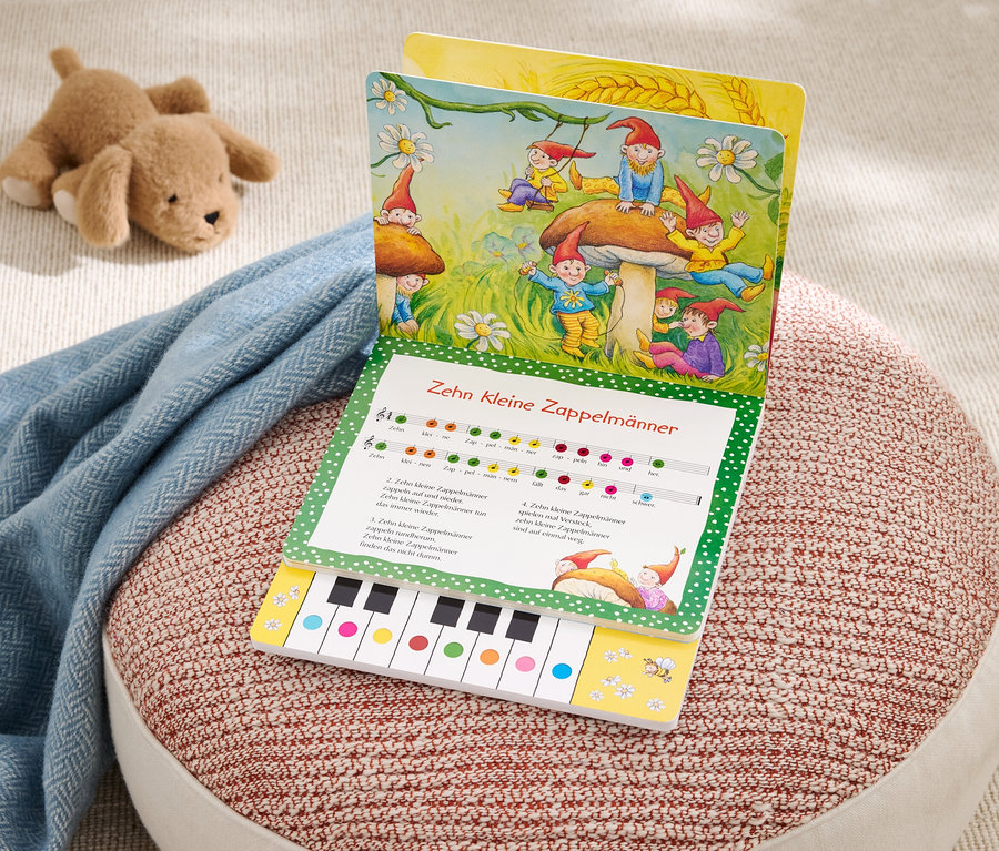 Ein Bilderbuch mit dem Titel "Zehn Kleine Zappelmaenner" liegt auf einem rot-weissen Pouf. Ein hellblaues Tuch und ein Stofftier liegen daneben.