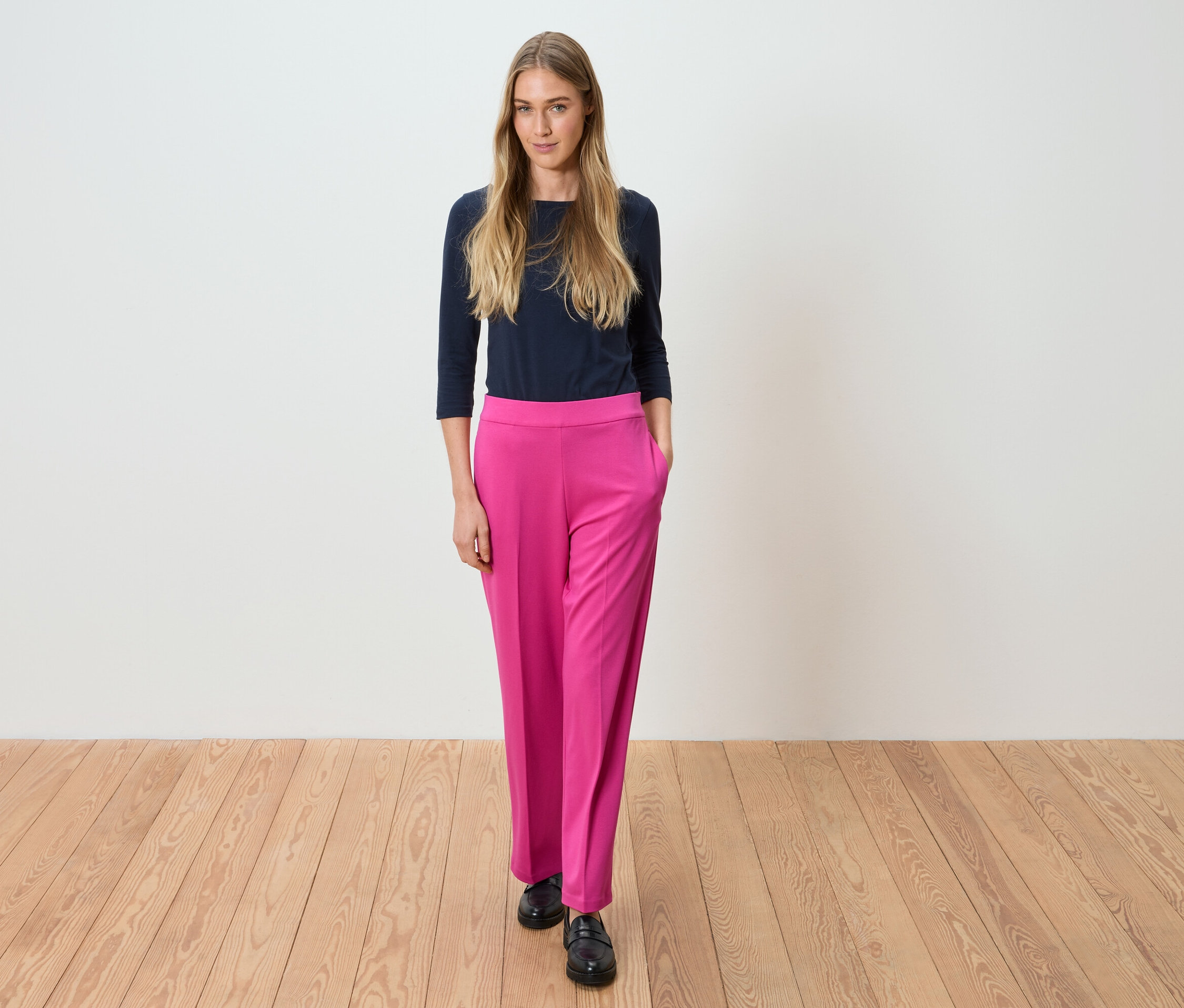 Frau steht in marineblauem Oberteil und pinker Hose vor einer weißen Wand.