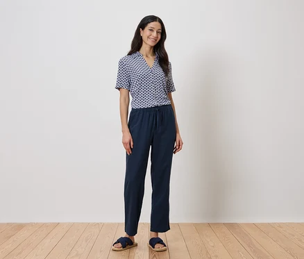 Eine Frau steht vor einer Wand und trägt ein blaues geometrisches Hemd und eine marineblaue Hose.