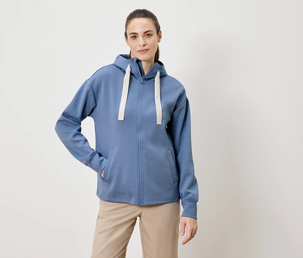 Frau mit Zopf trägt einen blauen Kapuzenpullover mit Reißverschluss und beigen Hosen.