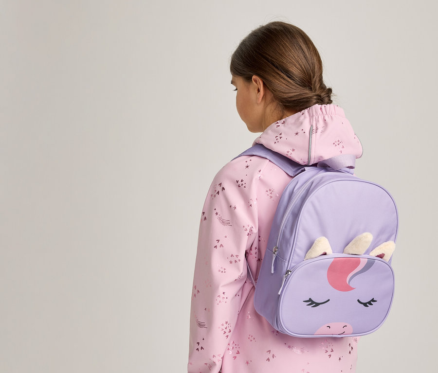 Ein junger Mensch von hinten gesehen, trägt eine rosa gemusterte Jacke und einen lila Einhorn-Rucksack mit Einhorn-Gesicht, Ohren und Horn.