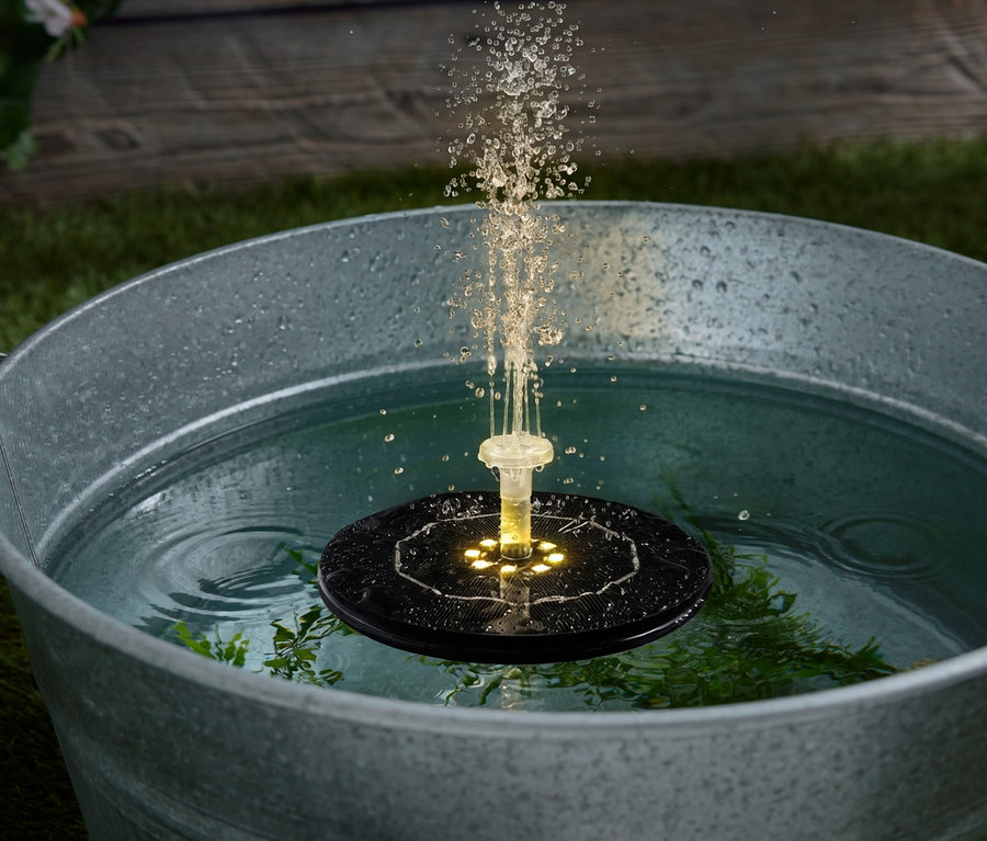 Eine silberne Wanne gefüllt mit Wasser, in der eine Solarbrunnenpumpe schwimmt und Wasser verspritzt.