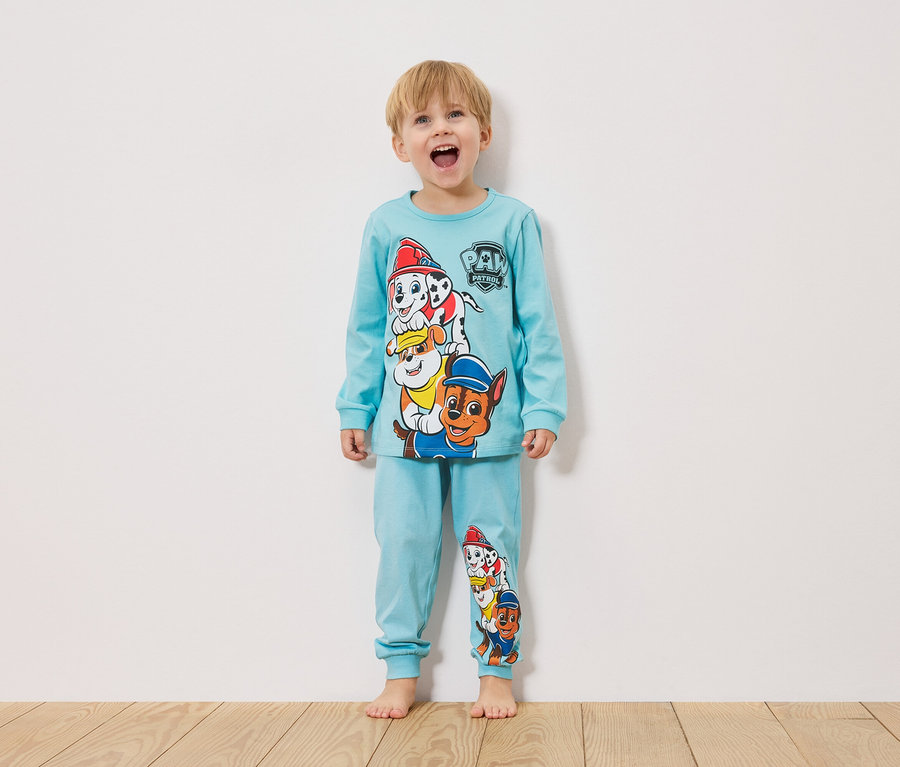 Ein kleiner Junge steht vor einer weißen Wand und trägt einen hellblauen Paw Patrol Pyjama.
