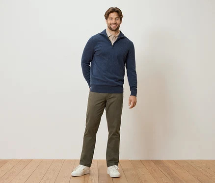 Ein Mann steht mit den Händen in den Hosentaschen vor einer Wand. Er trägt einen marineblauen Pullover, eine hellbraune Bluse, khakifarbene Hosen und weiße Turnschuhe.
