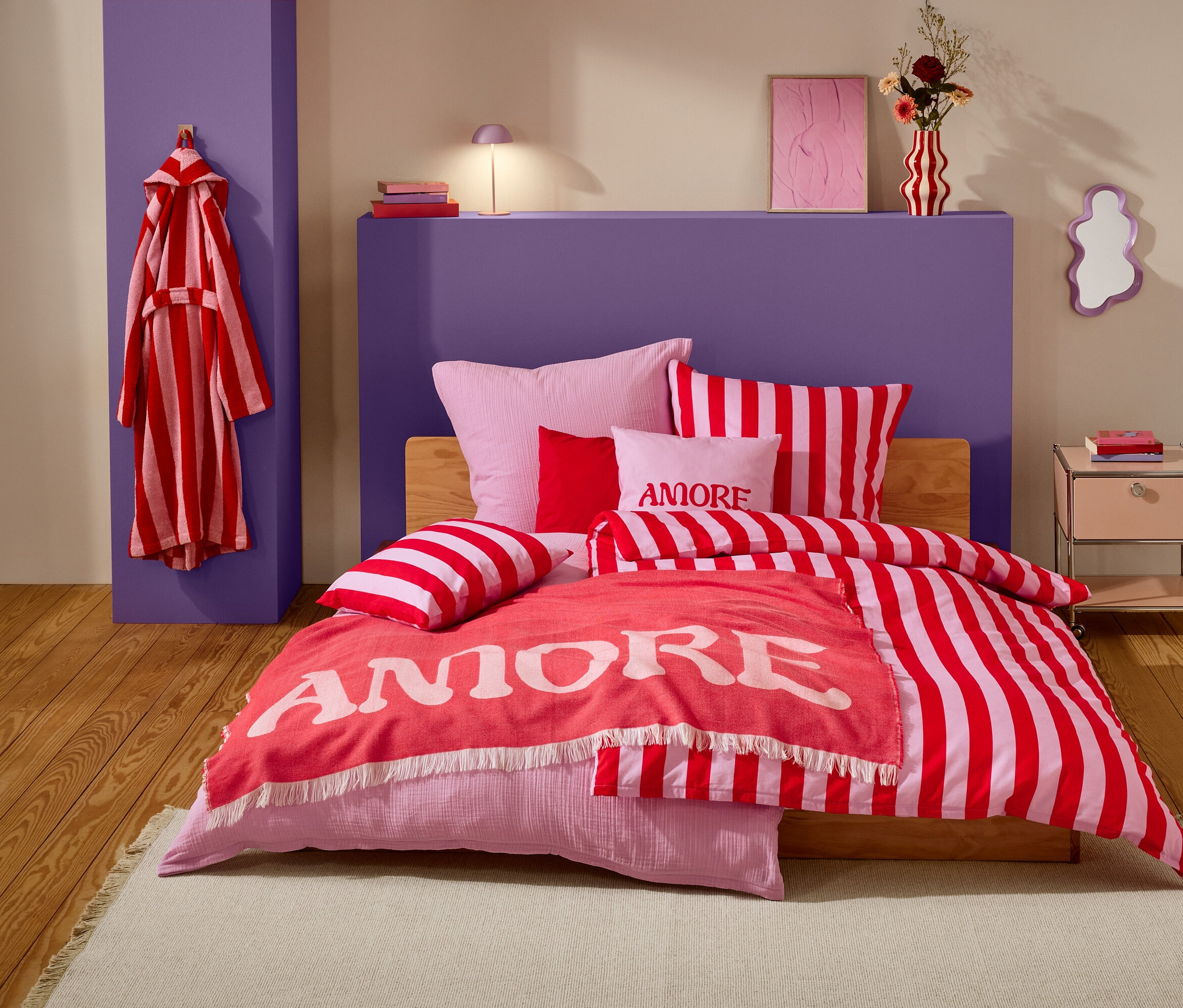 Ein Schlafzimmer mit einem Bett, das mit rot-weiß gestreifter Bettwäsche, rosa Kissen und einer korallenfarbenen Decke mit der Aufschrift "Amore" bedeckt ist. Ein rot-weiß gestreifter Bademantel hängt an einem violetten Pfeiler.