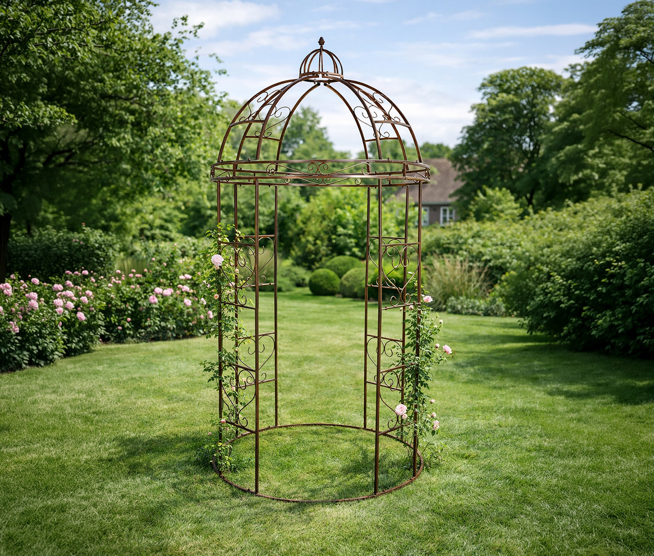 Auf Rasen steht ein bronzefarbener Rosenbogen, an dem Rosen hochranken. Im Hintergrund sind Bäume und ein Haus zu sehen.