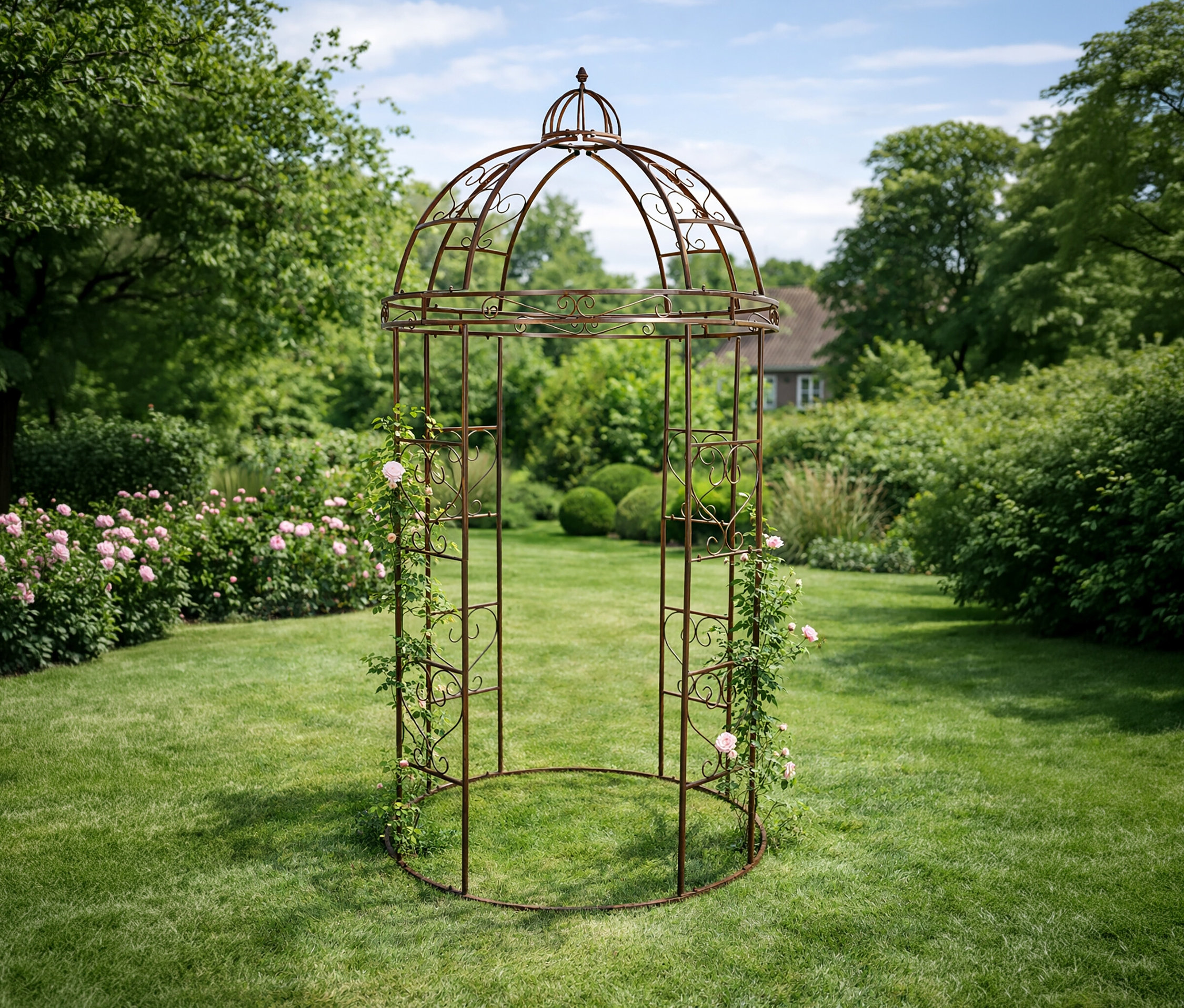 Auf Rasen steht ein bronzefarbener Rosenbogen, an dem Rosen hochranken. Im Hintergrund sind Bäume und ein Haus zu sehen.