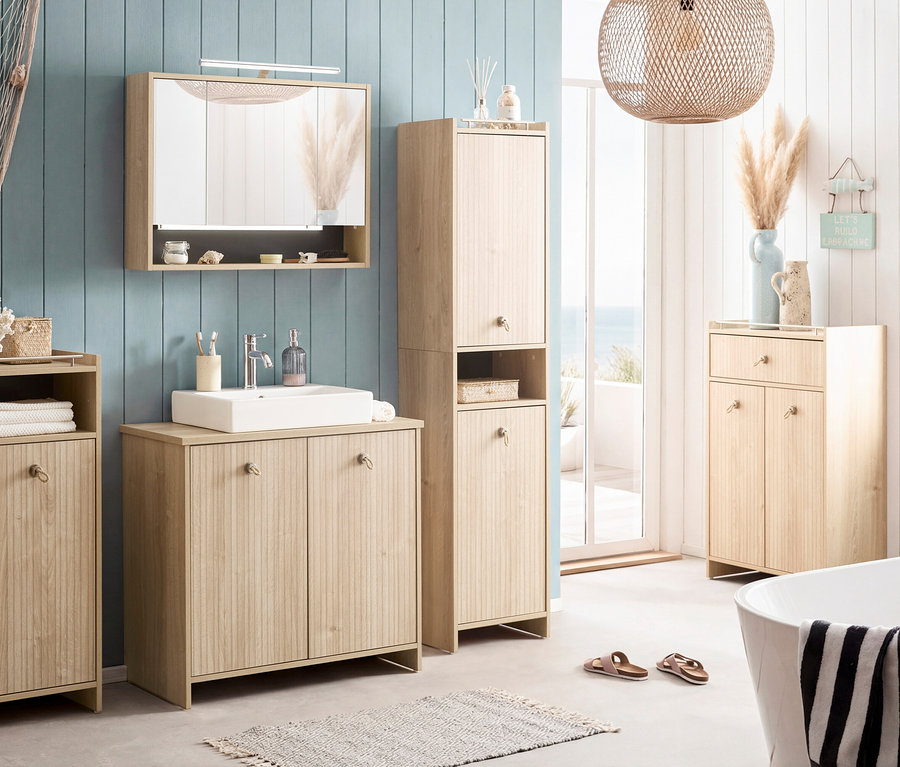 Badezimmer mit Waschtisch, Spiegelschrank, Hochschrank und Kommode in hellem Holz. Dekorationen im maritimen Stil und Badewanne im Vordergrund.