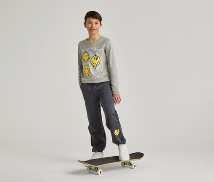 Ein Junge mit grauem Sweatshirt und Hose mit gelben Smileys steht auf einem Skateboard vor einem weißen Hintergrund.
