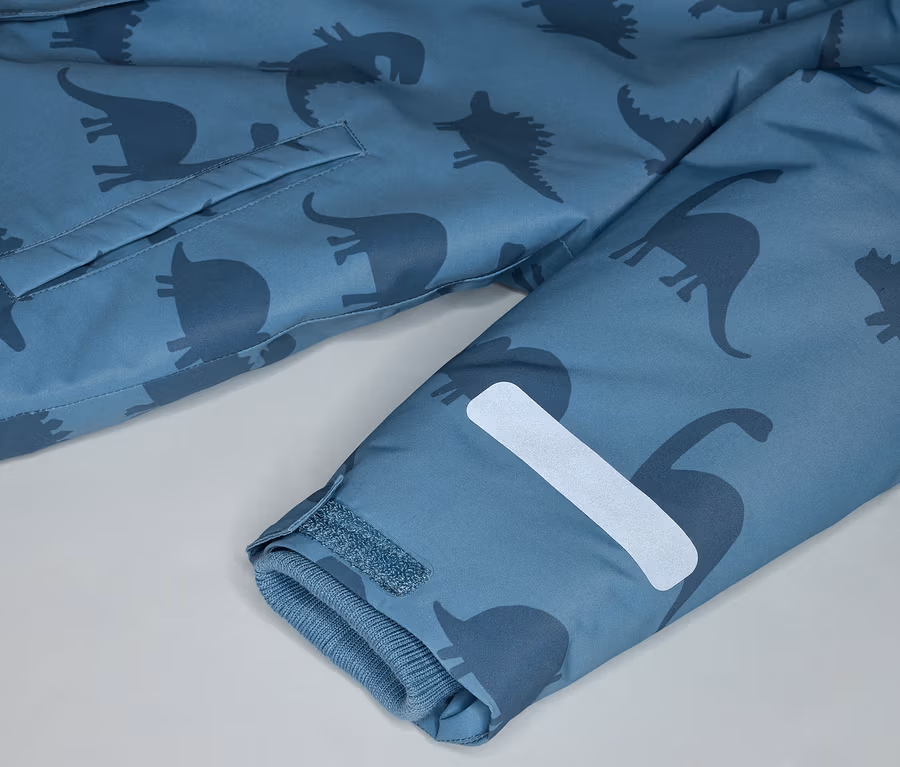 Nahaufnahme eines blauen Mantels mit Dinosaurier-Muster, einer Tasche und einem reflektierenden Streifen am Ärmel.