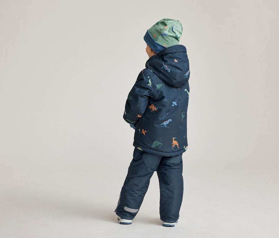 Ein Kind steht mit dem Rücken zur Kamera, es trägt eine marineblaue Jacke und Hose mit Dinosaurier-Motiven und eine grüne Mütze.