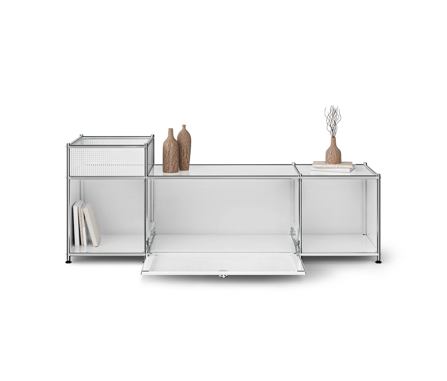 Weißes modulares Regal mit Chromrahmen, links ein perforierter Korb und Bücher, mittig eine offene Klapptür, rechts eine Vase mit Ästen und Bücher. Zwei braune Vasen stehen oben.