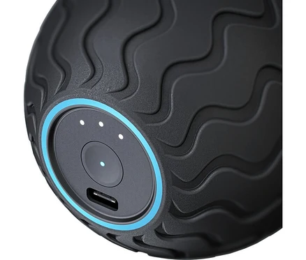Nahaufnahme eines schwarzen, texturierten Massagegeräts mit wellenförmigen Mustern. Es hat ein rundes Bedienfeld mit einem blauen Ring, einem zentralen Knopf, drei weißen Anzeigen (eine leuchtet) und einem USB-Anschluss.