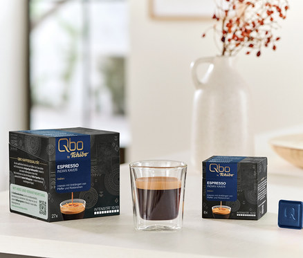 Auf einem Tisch stehen eine Tasse Espresso und Packungen Qbo Espresso INDIAN KAVERI mit 27 und 8 Kapseln.