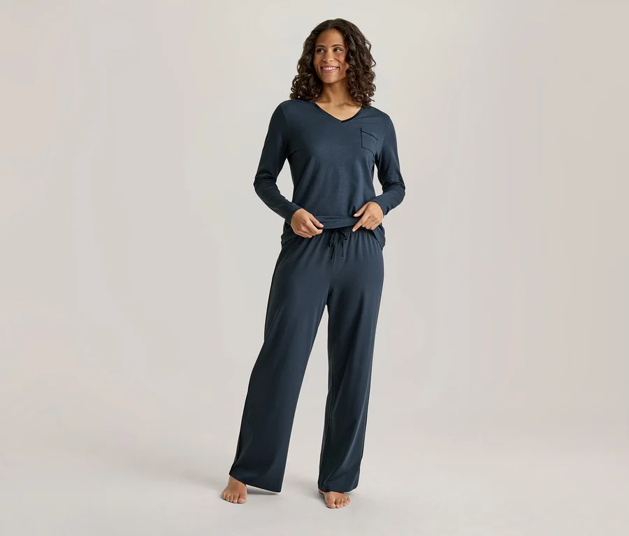Eine lächelnde Frau mit dunklem, lockigem Haar trägt ein dunkelblaues Langarm-V-Neck-Oberteil mit Tasche und eine passende weite Hose mit Kordelzug. Ihre Hände sind an ihrer Taille, und sie ist barfuß.