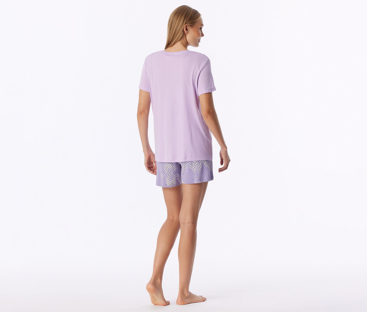 Frau steht mit dem Rücken zum Betrachter, trägt ein hellviolettes T-Shirt und gemusterte Shorts mit hellem geometrischem Muster. Sie ist barfuß.