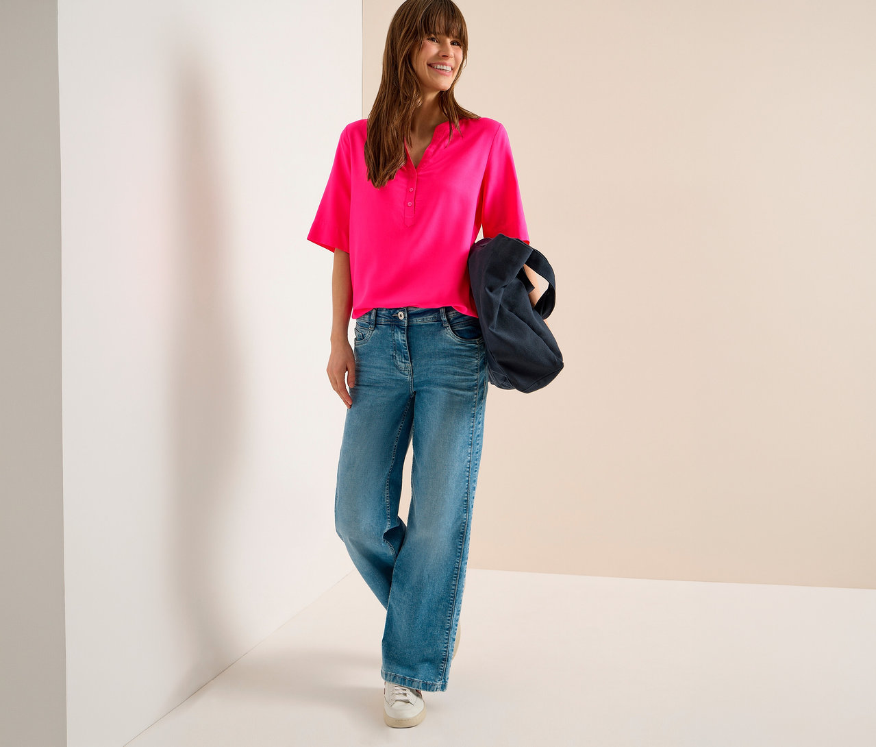 Lächelnde Frau in pinker CECIL Tunikabluse, blauen Wide-Leg-Jeans und weißen Sneakern, eine dunkle Tasche am Arm haltend.