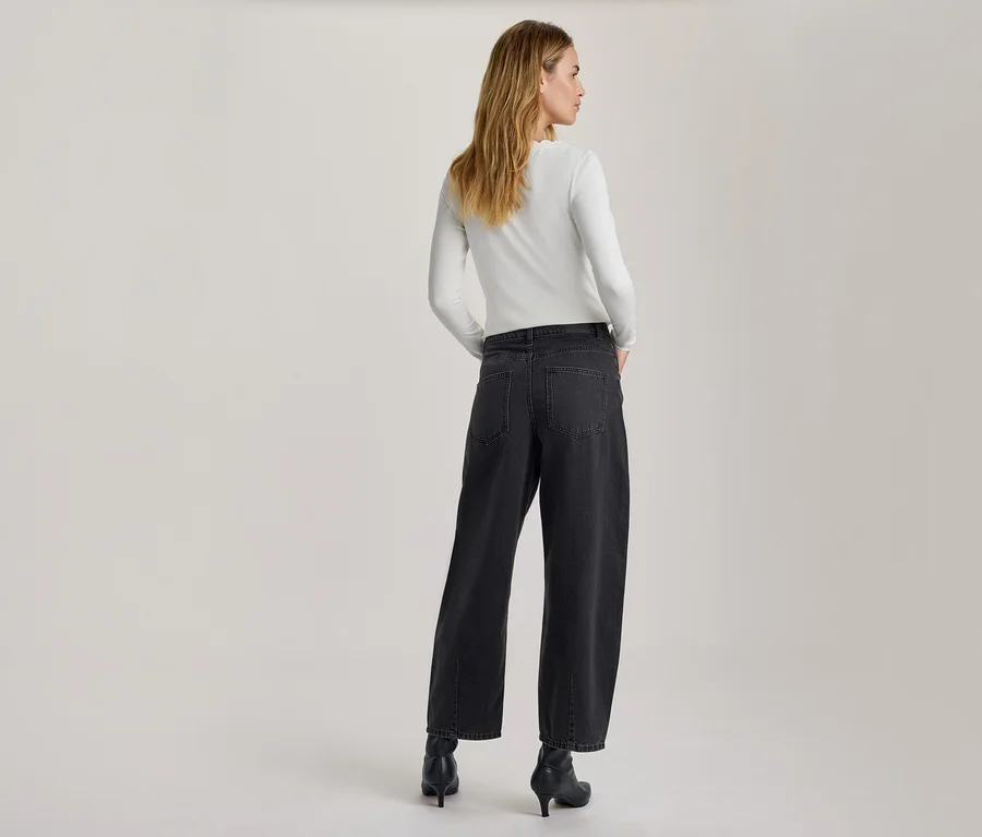 Frau von hinten, leicht zur Seite gedreht, trägt ein weißes Langarmshirt, eine dunkelgraue weite Jeans und schwarze Stiefeletten mit Absatz. Ihre Hände stecken in den Hosentaschen.