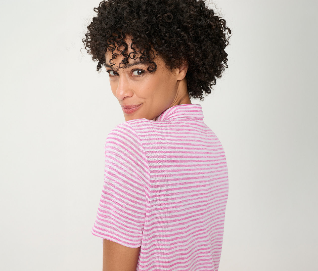 Frau mit lockigem Haar, die über ihre Schulter blickt und ein pink-weiß gestreiftes Olsen Poloshirt trägt.