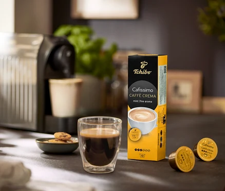 Auf einem Tisch stehen eine Kaffeemaschine, ein Glas Kaffee und eine Packung Cafissimo Caffè Crema mild - 120 Kapseln.