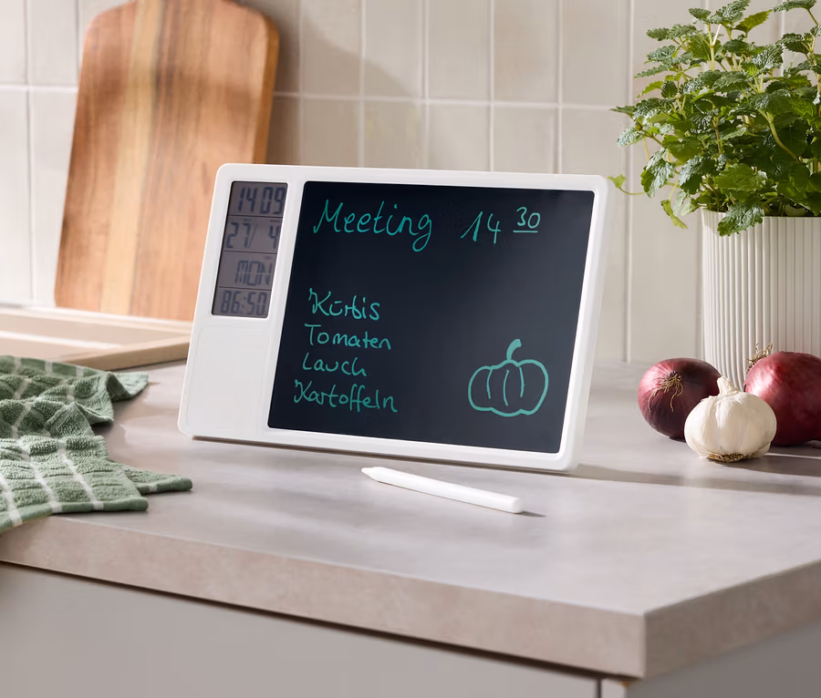 Weißes digitales Memoboard auf einer Küchenarbeitsplatte zeigt Meeting, Einkaufsliste, Uhrzeit, Datum und Temperatur. Daneben liegen Stift, grünes Tuch, Holzschneidebrett, Topfpflanze, rote Zwiebeln und Knoblauch.