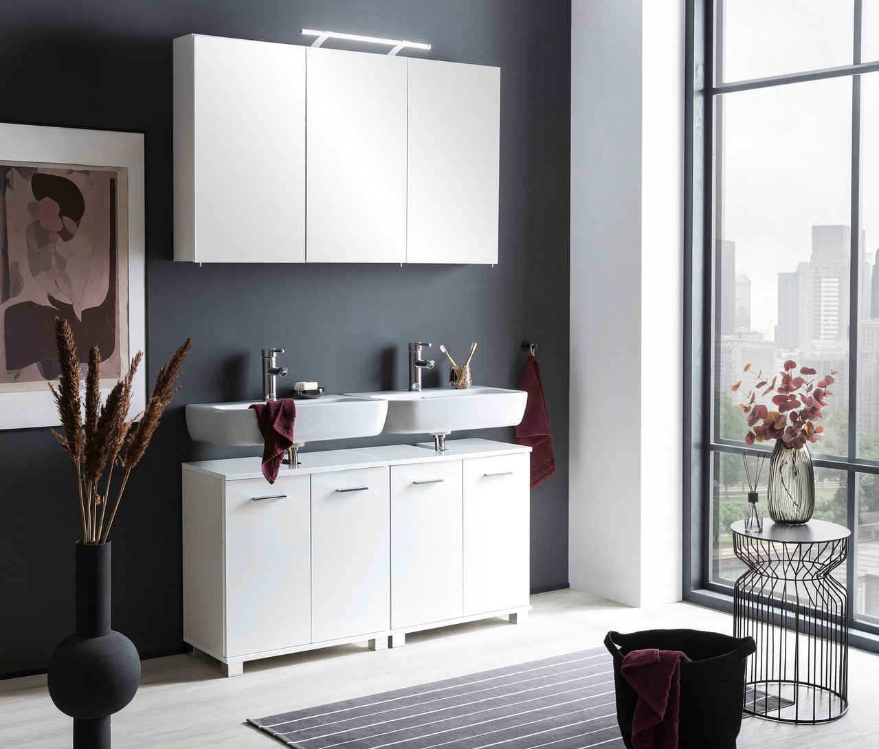 Modernes Badezimmer mit einem weißen Spiegelschrank »Dorina« mit Licht, 120 cm, über einem Doppelwaschbecken und einem weißen Unterschrank. Eine dunkelgraue Wand und ein großes Fenster mit Stadtblick sind zu sehen.