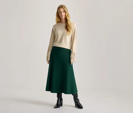 Frau mit langen blonden Haaren, die einen beigefarbenen Rundhalspullover, einen dunkelgrünen A-Linien-Midi-Rock und schwarze Stiefeletten trägt.