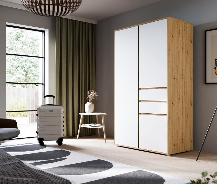 Ein modernes Schlafzimmer mit einem weiß-holzfarbenen Kleiderschrank, einem grauen Sessel und einem hellgrauen Koffer. Ein schwarz-weiß gemusterter Teppich liegt auf einem Holzboden, neben einem Fenster mit grünen Vorhängen und einer Stehlampe.
