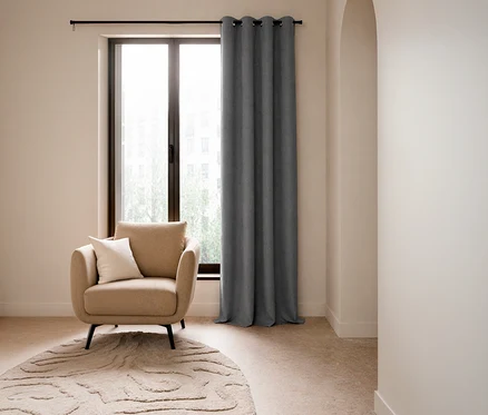 Ein beiger Sessel mit einem weißen Kissen steht vor einem großen Fenster, daneben hängt ein grauer SCHÖNER WOHNEN-Kollektion Verdunkelungsvorhang »OPACO«. Ein runder, gemusterter Teppich liegt auf dem Boden.