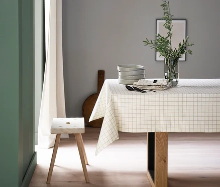 Ein Esstisch ist mit einer weiß-gold karierten SCHÖNER WOHNEN-Kollektion Tischdecke bedeckt. Auf dem Tisch stehen ein Stapel grauer Schüsseln, eine Vase mit grünen Zweigen und gefaltete Servietten mit Besteck. Ein Holzschemel steht neben dem Tisch.