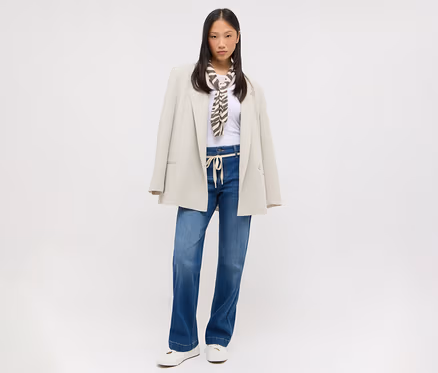 Junge asiatische Frau in weißem T-Shirt, blauen weiten Jeans mit hellem Kordelzug, beigem Oversize-Blazer über den Schultern und braun-weiß gemustertem Schal. Sie trägt weiße flache Schuhe.