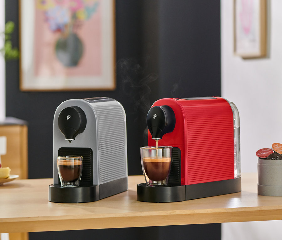 Zwei Kaffeemaschinen, eine graue und eine rote, stehen auf einem Holztisch. Die rote Maschine brüht Kaffee in eine Glastasse, während die graue Maschine eine volle Tasse hat. Ein Behälter mit Kaffeekapseln und ein Stück Himbeerkuchen sind ebenfalls auf dem Tisch.