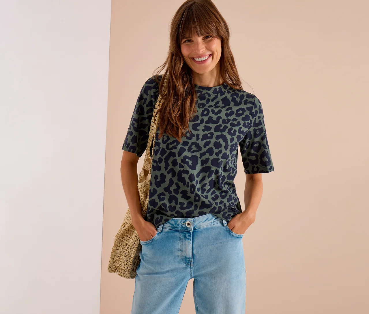Lächelnde Frau mit langen Haaren trägt ein dunkelgrünes CECIL Jerseyshirt mit Leopardenmuster, helle Jeans und eine gewebte Tasche.