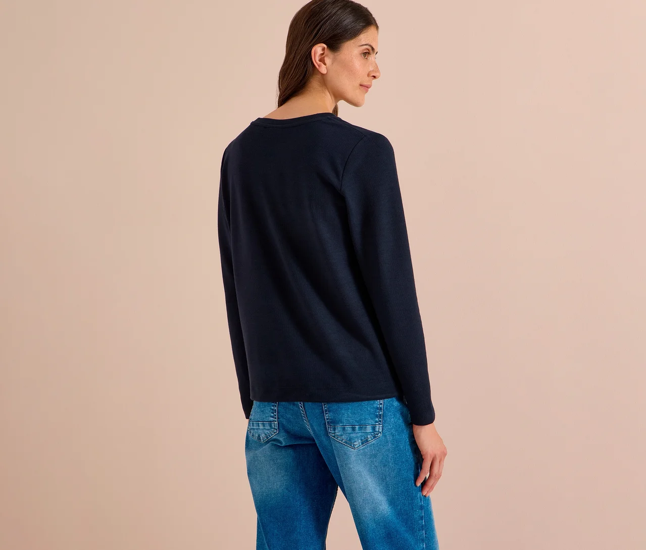 Frau von hinten, leicht nach rechts gedreht, trägt ein dunkelblaues CECIL Shirt und blaue Jeans.