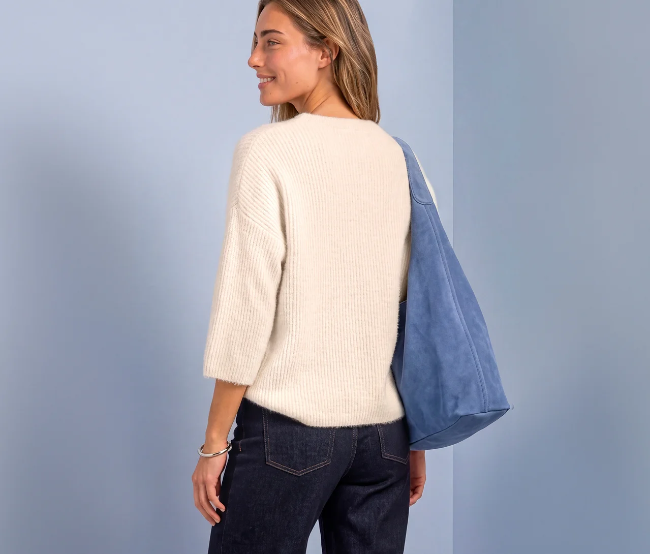 Frau von hinten in einem cremefarbenen Street One Strickpullover, dunklen Jeans und blauer Wildledertasche, blickt über die Schulter.