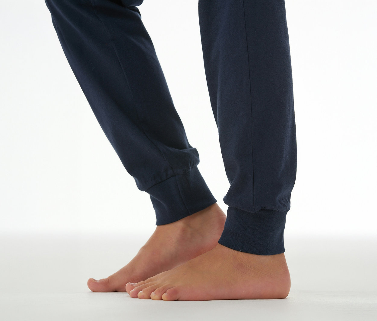 Unterschenkel und nackte Füße einer Person, die eine dunkelblaue SCHIESSER Kinder-Pyjamahose mit Bündchen trägt und auf einem weißen Untergrund steht.