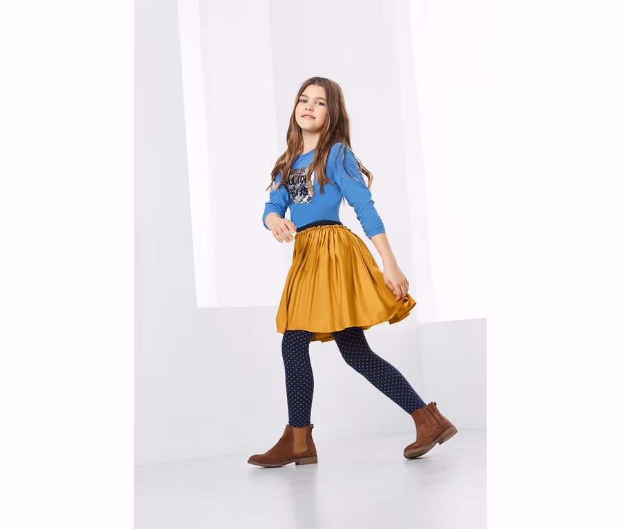 Mädchen geht in blauem Shirt, Curry-Rock, gemusterter Leggings und braunen Stiefeln.