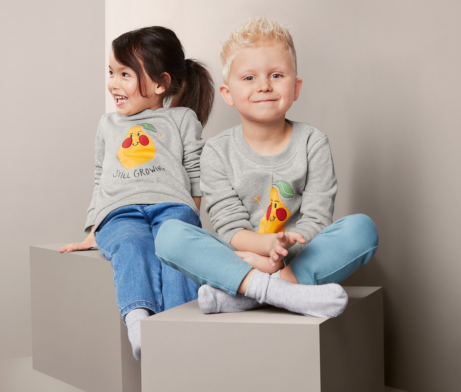 Zwei Kinder sitzen auf grauen Blöcken und tragen graue Sweatpullover mit Birnenmotiven.
