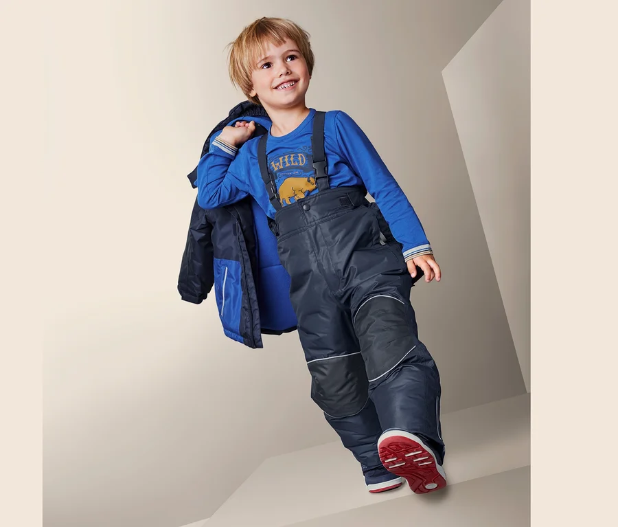 Junge in Schneehose mit recyceltem Material, blauem Shirt und Jacke über der Schulter geht Treppe runter.