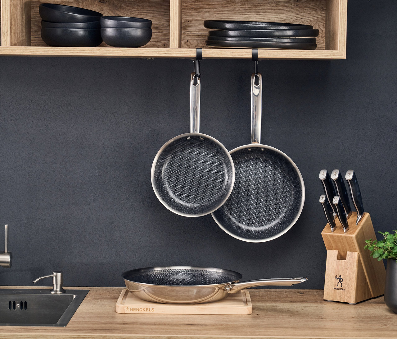 Drei Bratpfannen des Henckels by ZWILLING »Valley« Bratpfannensets in einer Küche: zwei hängen unter einem Holzregal, eine steht auf einem Holzschneidebrett neben einem Spülbecken und einem Messerblock.