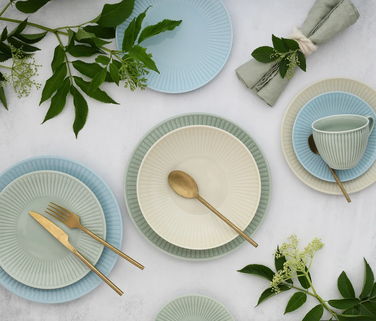 Ein creaTable Kombiservice »Valencia Pastell« auf einer hellen Fläche, bestehend aus gerippten Tellern, Schalen und Tassen in Pastellfarben: Hellblau, Hellgrün und Creme. Goldenes Besteck, eine grüne Serviette und grüne Blätter mit Blüten schmücken die Anordnung.