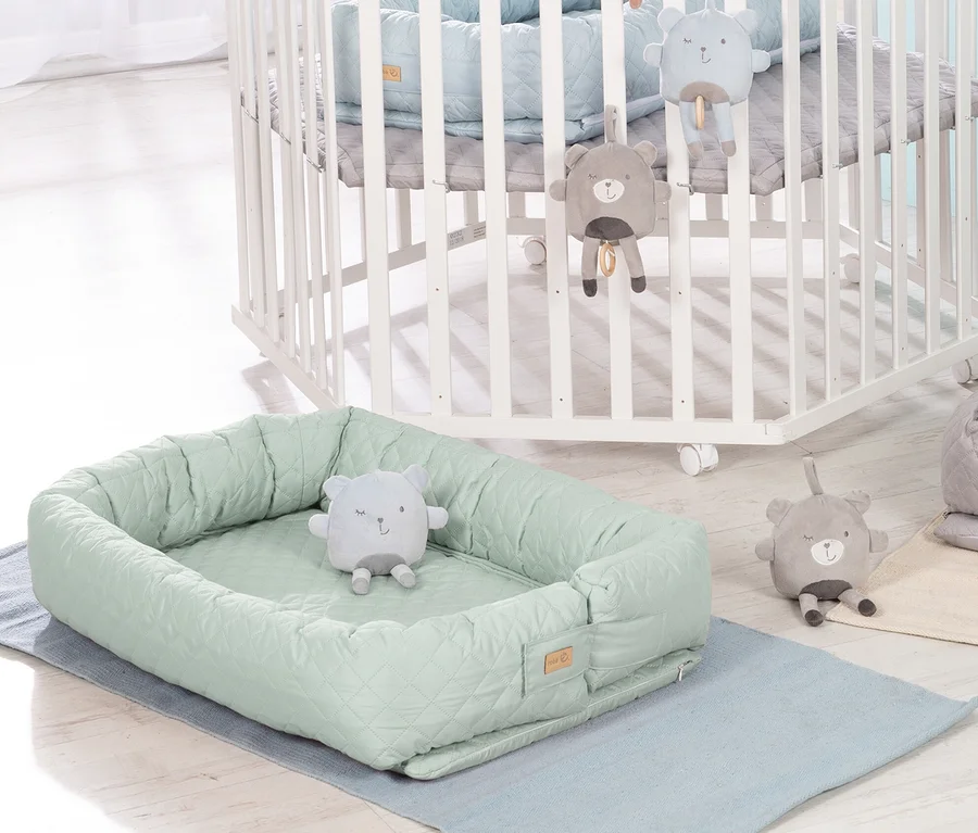 Grüne Roba 3-in-1 Babylounge mit Teddybär-Spielzeug liegt auf einer blauen Unterlage neben einem weißen Kinderbett.