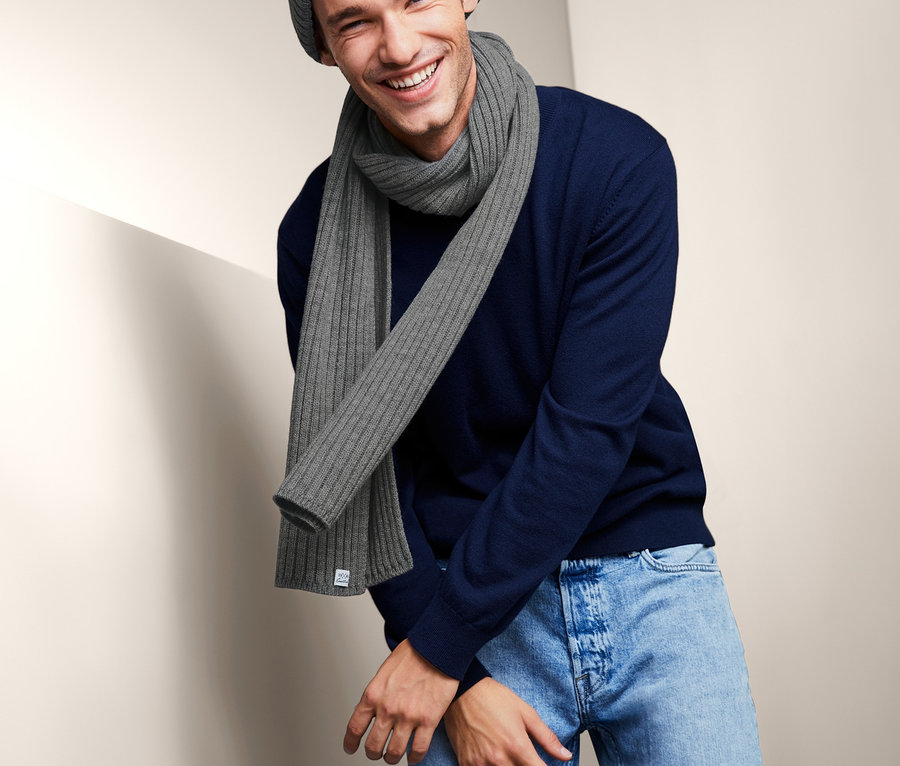 Mann lächelt und trägt einen Strickschal, eine Strickmütze und einen blauen Pullover.